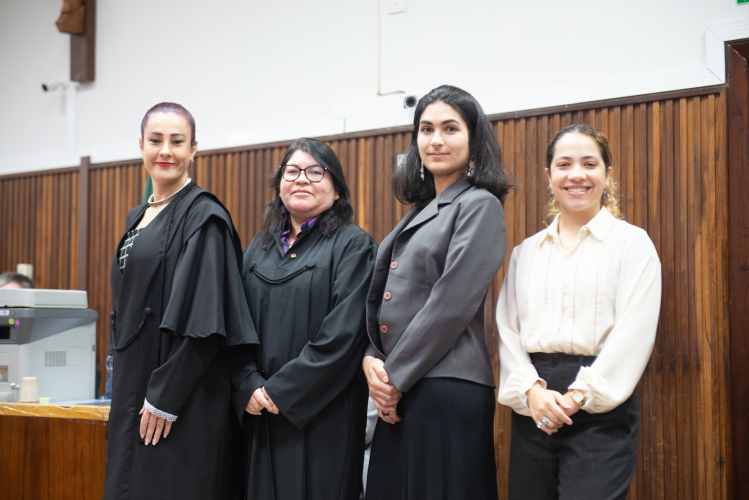Quatro mulheres posam em um tribunal. Duas usam togas pretas, sendo uma com cabelo preso e a outra com óculos. As outras duas estão de traje social: uma com blazer cinza e cabelo solto, e outra com camisa clara, calça preta e cabelo preso. Ao fundo, parede de madeira com detalhes claros.