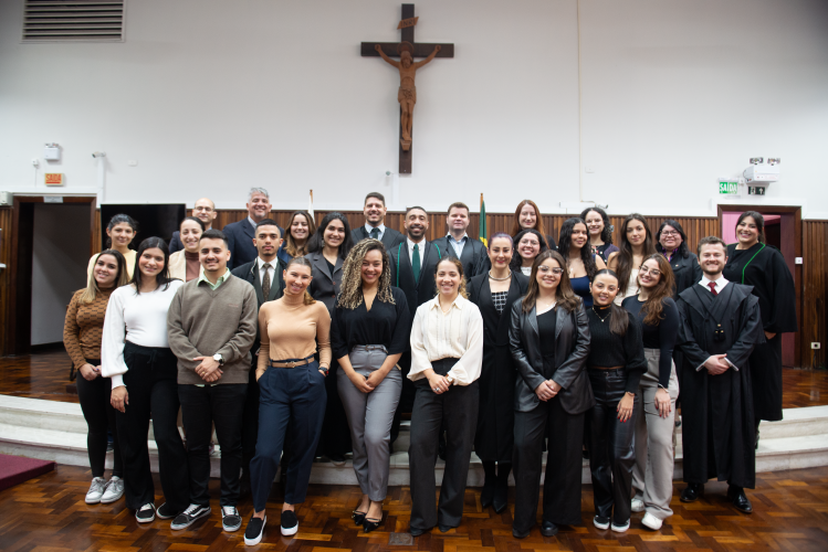 Foto de grupo com cerca de 30 pessoas reunidas no tribunal. Alguns vestem togas pretas, enquanto outros estão em trajes sociais variados. Todos sorriem, posicionados em três fileiras. Ao fundo, parede de madeira, crucifixo grande e bandeiras.