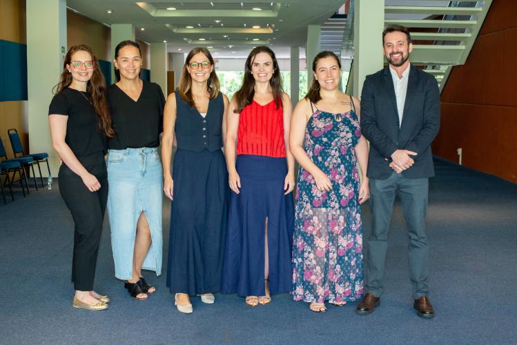 Cinco mulheres e um homem pousam para a foto sorrindo. Eles estão com roupas levemente formais e estão em um ambiente iluminado no corredor da Universidade.