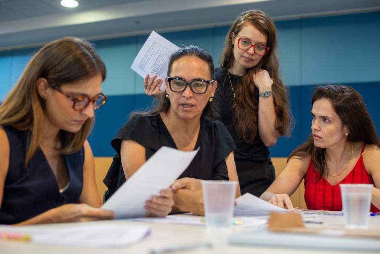 A imagem retrata um momento de análise e discussão durante a prova oral do V Concurso para defensores e defensoras. Quatro mulheres estão reunidas em uma mesa, examinando documentos e debatendo com expressões concentradas e sérias.