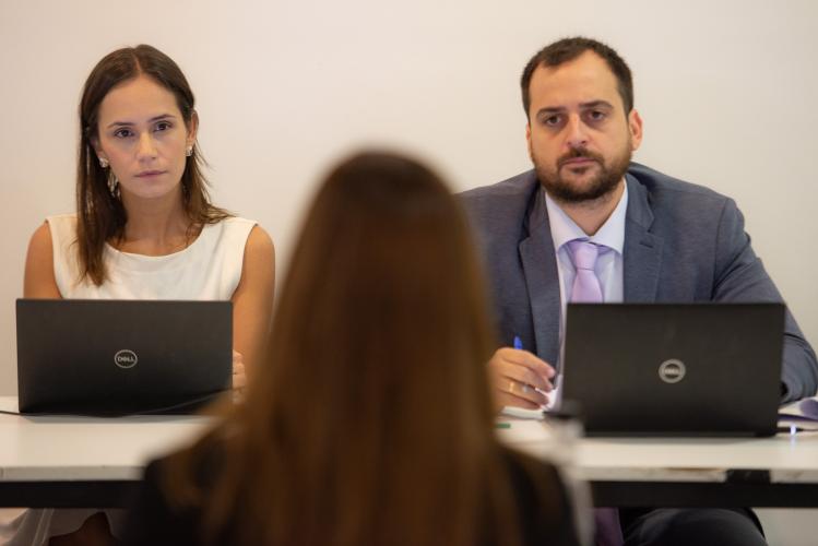 Imagem de dois defensores, um homem e uma mulher, que olham atentamente para a candidata que está desfocada de costas. Eles olham seriamente enquanto estão com o computador na frente.