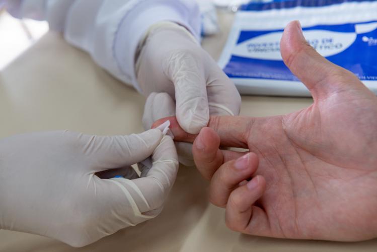 Foto em que paciente realizada coleta de DNA com um profissional de saúde. A imagem mostra apenas a mão direita do paciente, utilizada para coleta, e as mãos do profissional, utilizando luvas para o exame.