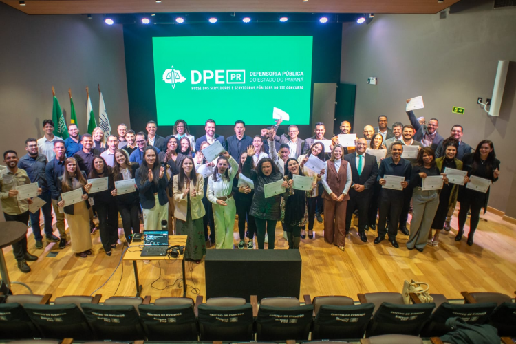 Imagem com foto dos 56 servidores e servidoras aprovadas no III concurso da DPE-PR, em um palco, à frente de uma tela com a logo da Defensoria Pública.