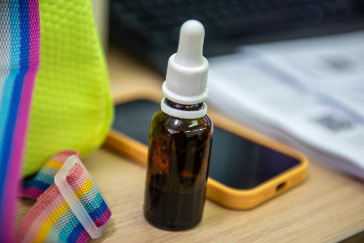 Imagem de um frasco de conta gotas comumente usado para guardar Canabinol sobre uma mesa onde se vê detalhe de uma bolsa e de um celular.