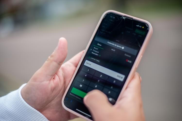 Imagem em close de um celular nas mãos de um apostador. Tela mostra uma aposta no jogo Brasil x Paraguai feita em plataforma de bets. Ao fundo a rua desfocada.