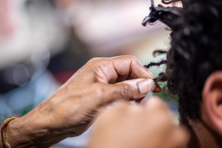 Imagem com foto em que cabeleireiro realiza tranças nos cabelos escuros de uma pessoa negra.