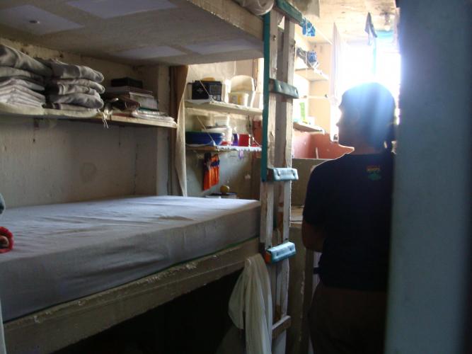 Imagem com foto de ambiente interno de uma cela, com uma mulher ao fundo, de costas, com o rosto virado para a janela, por onde entra a luz do sol.