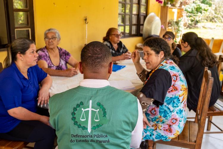 Imagem que mostra um atendimento da DPE-PR em área rural. A foto, tirada por trás de um homem, mostra um encontro entre um representante da Defensoria Pública, identificado por seu colete verde com o logo da instituição, e um grupo de mulheres. Todos estão sentados ao redor de uma mesa branca em uma área de varanda com uma parede amarela ao fundo, e a cena captura um momento de diálogo, com uma das mulheres, de blusa floral colorida, falando ativamente para o grupo.