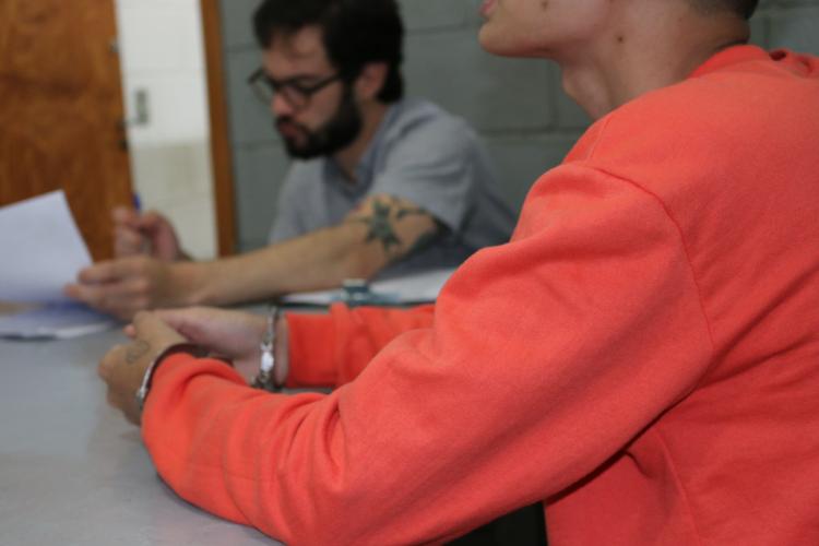 Em primeiro plano, uma pessoa vestindo um moletom laranja está sentada em uma mesa, com os pulsos unidos por algemas. Do outro lado da mesa, em segundo plano, um homem de barba e óculos, vestindo uma camiseta cinza, lê um documento. A cena se passa em uma sala com parede de blocos de concreto.
