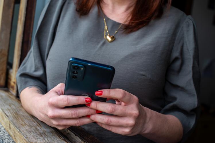Imagem em close-up de uma mulher que segura um celular com as duas mãos. Ela veste uma blusa cinza-escura e usa um colar dourado com um pingente de coração. Suas unhas, pintadas de um vermelho vivo, destacam-se enquanto ela manuseia o aparelho de cor azul-escura.