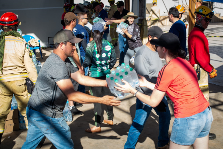 Imagem que mostra o recebimento de doações em Rio Bonito do Iguaçu. Em primeiro plano, um homem de camiseta cinza entrega um fardo grande de garrafas de água para uma mulher de camiseta vermelha. Ao fundo, voluntários e pessoas com uniformes de resgate participam da distribuição.