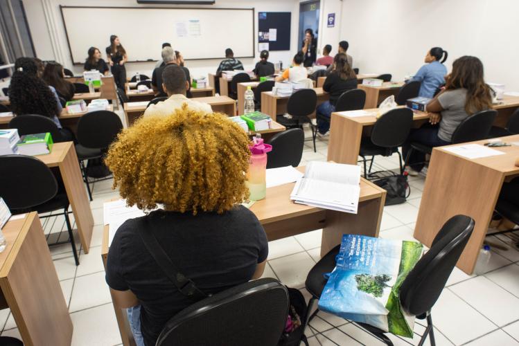 A imagem mostra uma sala de aula com alunos adultos sentados em carteiras de madeira voltadas para um quadro branco ao fundo. Em primeiro plano, vista de costas, destaca-se uma pessoa com cabelos crespos e volumosos em tom loiro. Sobre as mesas, há cadernos e materiais de estudo.