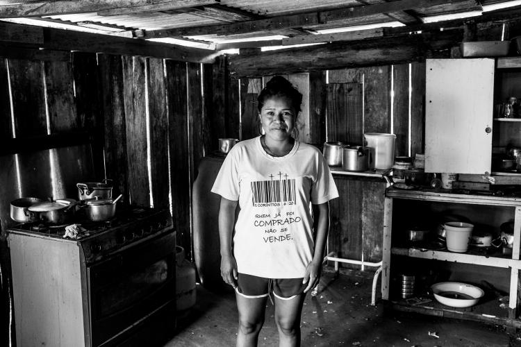 Fotografia em preto e branco de uma mulher em pé no centro de uma cozinha rústica com paredes de madeira. Ela veste uma camiseta com um código de barras e a frase "Quem já foi comprado não se vende", cercada por um fogão com panelas à esquerda e prateleiras simples com utensílios à direita.