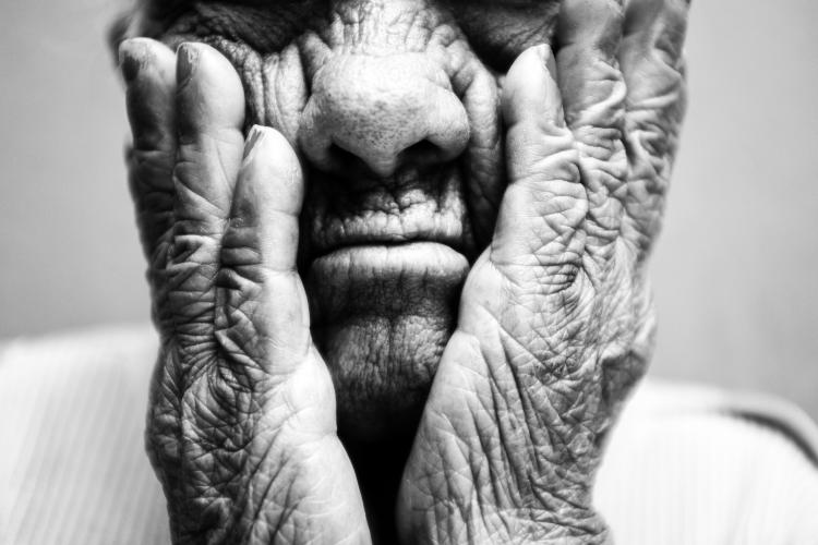 Fotografia em preto e branco com foco em um close-up do rosto e das mãos de uma pessoa idosa. As mãos, com rugas profundas e marcantes, apoiam as bochechas e emolduram o nariz e a boca de lábios cerrados, destacando intensamente a textura da pele.