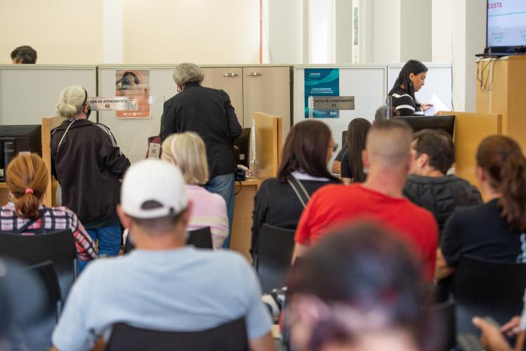 Vista de trás de uma sala de espera cheia, onde diversas pessoas estão sentadas de costas em cadeiras pretas. No fundo, balcões de atendimento de madeira e divisórias brancas têm placas com os dizeres "MESA DE ATENDIMENTO". Uma funcionária é visível atrás de um balcão manuseando documentos.