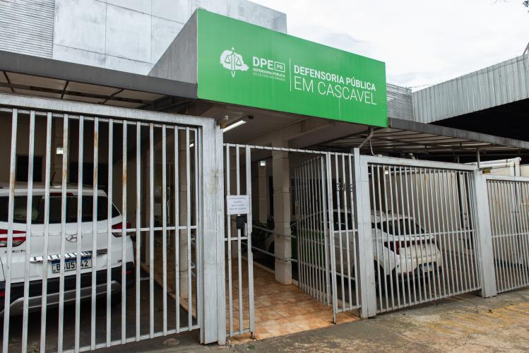 Imagem que mostra a fachada da sede da Defensoria Pública em Cascavel. O local apresenta uma grande placa verde com o logotipo da DPE-PR na parte superior e um portão de grades metálicas brancas na entrada, abrigando dois carros brancos estacionados na garagem.