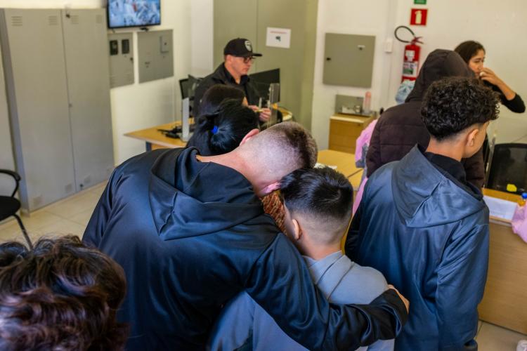 A foto, tirada de um ângulo superior, mostra pessoas de costas em uma sala de atendimento. Em primeiro plano, um homem de moletom preto abraça um menino pelos ombros. Ao fundo, um homem de boné e óculos atende o público atrás de um balcão, em um ambiente com armários cinzas e um extintor de incêndio na parede.