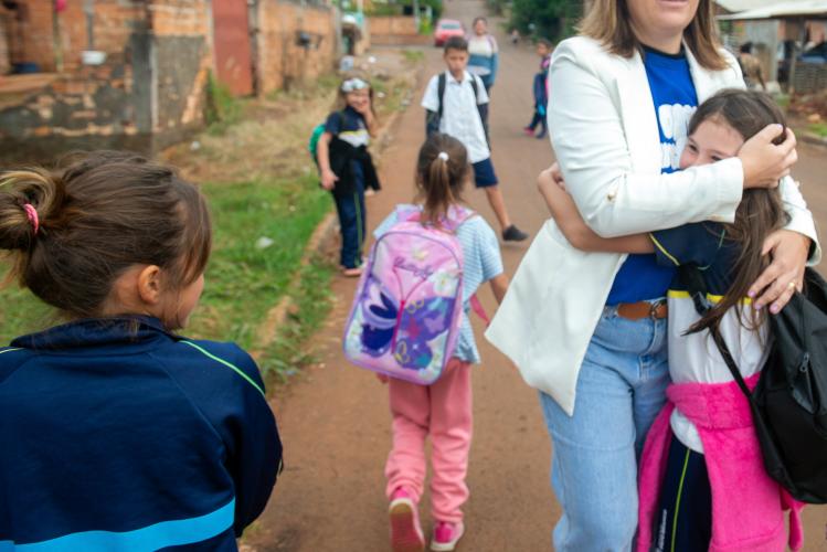 Fotografia em uma rua de terra onde várias crianças com uniformes escolares caminham. Em primeiro plano à direita, uma mulher de blazer branco abraça afetuosamente uma menina, enquanto outra garota à esquerda, vista de costas, observa a cena. Ao fundo, outras crianças seguem pela via.