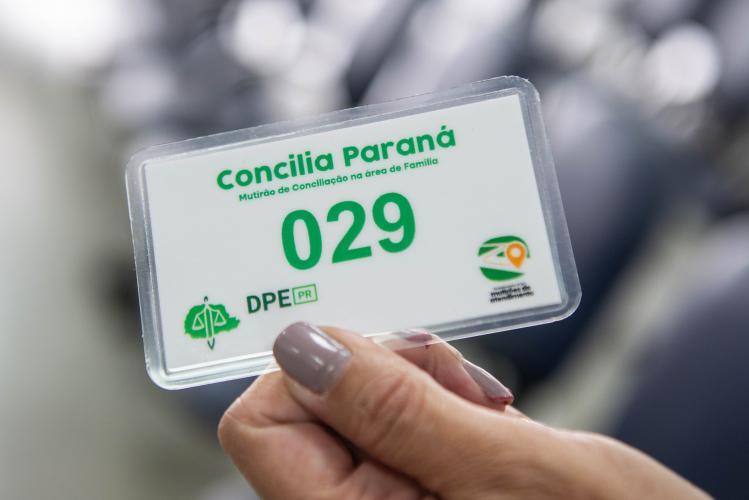 Fotografia em close de uma mão segurando uma senha plastificada de fundo branco. Em letras verdes, lê-se "Concilia Paraná - Mutirão de Conciliação na área de Família" com o número "029" em grande destaque no centro, acompanhado dos logotipos da DPE-PR e do mutirão na base.