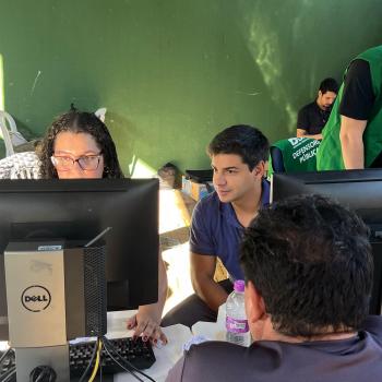 Imagem mostra três servidores utilizando computadores durante os atendimentos do mutirão. De costas em primeiro plano se vê um assistido.