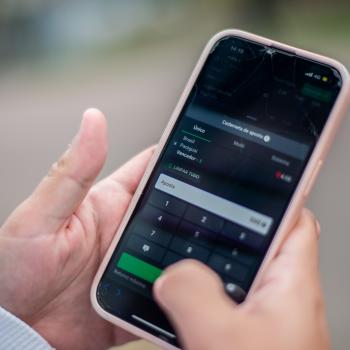 Imagem em close de um celular nas mãos de um apostador. Tela mostra uma aposta no jogo Brasil x Paraguai feita em plataforma de bets. Ao fundo a rua desfocada. 