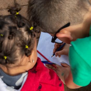 Duas crianças pequenas, um menino de verde e uma menina de vermelho, fazem atividade com lápis de cor. 