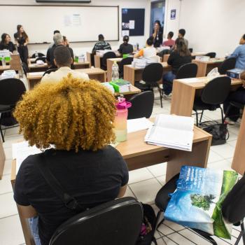 A imagem mostra uma sala de aula com alunos adultos sentados em carteiras de madeira voltadas para um quadro branco ao fundo. Em primeiro plano, vista de costas, destaca-se uma pessoa com cabelos crespos e volumosos em tom loiro. Sobre as mesas, há cadernos e materiais de estudo.