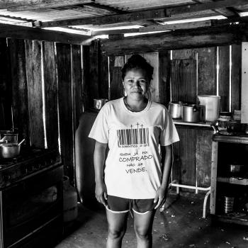 Fotografia em preto e branco de uma mulher em pé no centro de uma cozinha rústica com paredes de madeira. Ela veste uma camiseta com um código de barras e a frase "Quem já foi comprado não se vende", cercada por um fogão com panelas à esquerda e prateleiras simples com utensílios à direita.