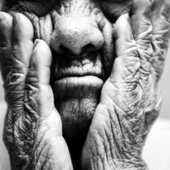 Fotografia em preto e branco com foco em um close-up do rosto e das mãos de uma pessoa idosa. As mãos, com rugas profundas e marcantes, apoiam as bochechas e emolduram o nariz e a boca de lábios cerrados, destacando intensamente a textura da pele.