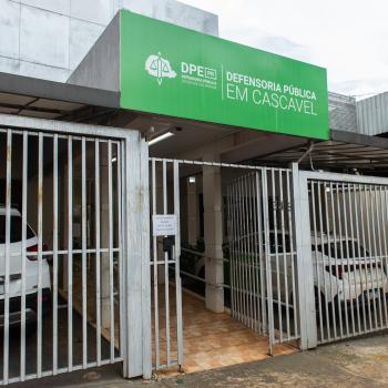 Imagem que mostra a fachada da sede da Defensoria Pública em Cascavel. O local apresenta uma grande placa verde com o logotipo da DPE-PR na parte superior e um portão de grades metálicas brancas na entrada, abrigando dois carros brancos estacionados na garagem.