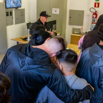 A foto, tirada de um ângulo superior, mostra pessoas de costas em uma sala de atendimento. Em primeiro plano, um homem de moletom preto abraça um menino pelos ombros. Ao fundo, um homem de boné e óculos atende o público atrás de um balcão, em um ambiente com armários cinzas e um extintor de incêndio na parede.
