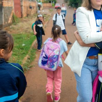 Fotografia em uma rua de terra onde várias crianças com uniformes escolares caminham. Em primeiro plano à direita, uma mulher de blazer branco abraça afetuosamente uma menina, enquanto outra garota à esquerda, vista de costas, observa a cena. Ao fundo, outras crianças seguem pela via.