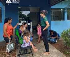 Foto de uma servidora branca com calça preta e camiseta da instituição na cor verde, conversando com usuárias da defensoria que esperam sentadas em cadeiras do lado de fora do prédio do CRAS. O prédio é de cor azul, em frente estão três cadeiras verdes e uma preta, onde estão sentadas duas crianças acompanhadas de suas mães, sendo elas uma mulher negra e duas mulheres brancas. Mais ao lado, uma criança branca aguarda ao lado da porta e, agachado também ao lado da porta, um homem mexe no celular.