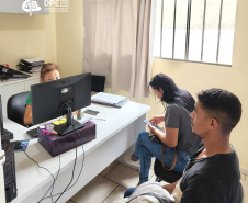 Foto de uma servidora amarela com a camiseta da instituição mexendo em um computador enquanto atende dois usuários, sendo eles um homem negro de camisa preta e branca e calça jeans, e uma mulher branca com calça jeans e camiseta cinza mexendo no celular.