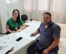 Imagem de uma servidora da defensoria com pele branca e camiseta da instituição na cor verde, com os braços apoiados em uma mesa branca. Ela atende um usuário branco com camiseta e calça em tons de azul. Ele segura uma caneta para assinar alguns papeis que estão sobre a mesa.