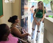 Imagem de duas mulheres negras usuárias da defensoria sentadas na parte interna do prédio do CRAS sendo atendidas por uma servidora branca que usa a camiseta da instituição e calça jeans.