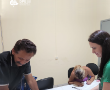 Imagem de uma servidora da Defensoria com a camisa da instituição em verde entregando documento para um homem branco e, ao fundo, uma mullher branca assina alguns papeis em uma mesa branca.