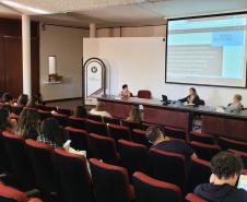 Imagem que mostra uma das palestras do evento. Três mulheres fazem uma apresentação, sentadas atrás de uma mesa, para uma plateia formada por várias pessoas que as escutam com atenção.