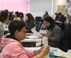 Imagem que mostra uma pessoa sendo atendida em Curitiba, ela está segurando um papel e olhando para ele. Ao fundo, outras pessoas aguardam atendimento.