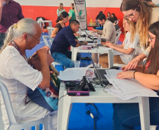 Imagem que mostra uma mulher sendo atendida por profissionais da DPE-PR no mutirão em Paranaguá. Elas estão em uma mesa com cadeiras plásticas, brancas, em um ginásio. Ao fundo, mais mesas de atendimento e pessoas sendo atendidas.