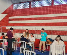 Imagem que mostra o atendimento no mutirão em Paranaguá, com três mesas com cadeiras plásticas, brancas, em um ginásio. Nas três estão pessoas sendo atendidas.