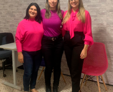 Na foto estão pousando as secretárias de atendimento Daiane Cristiane Pereira dos Santos e Dioneia dos Santos Modesto, e a defensora pública de Paranaguá Ingrid Lima Vieira, nesta ordem da esquerda para a direitas. As três estão sorrindo, com roupas cor de rosa e roxa. 