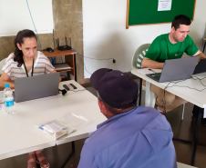 Imagem que mostra um atendimento no mutirão. Uma mulher atende um homem em uma mesa. Na mesa ao lado, o defensor público Matheus Munhoz também faz um atendimento.