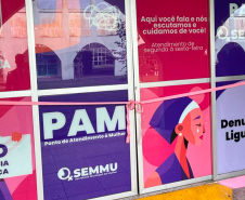Fotografia da porta de entrada do PAM. A porta é de vidro com adesivo de artes gráficas roxa e rosa. Na frente da porta está uma fita rosa, fazendo alusão à inauguração.