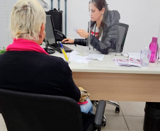 Imagem que mostra um atendimento do mutirão. A atendente da Defensoria analisa um papel e, em cima da mesa onde ela trabalha, estão cartilhas com o tema do mutirão.