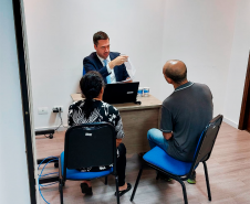 Imagem que mostra o defensor Matheus Munhoz atendendo um homem e uma mulher em uma mesa de atendimento com um notebook em cima. Eles estão sentados de frente para o defensor público.
