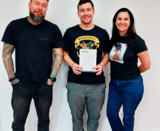 Imagem que mostra a usuária da DPE-PR Marty Cristine Maceno com seu filho e o pai dele, Clayton Teles dos Santos. Os três estão em pé, o filho ao centro, segurando uma pasta com o resultado do DNA.