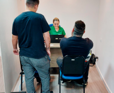 Imagem da assessora Bruna Abdalla atendendo dois usuários da DPE-PR, um deles está de pé e o outro está sentado. Ela está sentada de frente para eles e sorri.