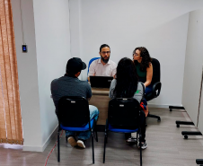 Imagem que mostra o defensor Fernando Redede e a psicóloga Lethicia Gaidarji Silva atendendo a um casal de usuários da DPE-PR, todos estão sentados em volta de uma mesa.