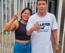 Imagem onde mostra a mãe com seu filho mais velho, ambos sorriem e posam para a foto em frente a um portão branco.